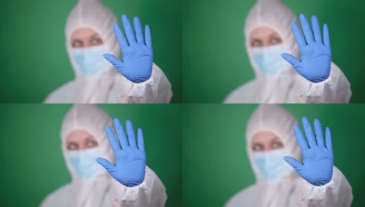 年轻的女医生穿着白色的鼠疫防护服，戴着蓝色的橡胶手套，在绿色的背景下显示停止的手势高清在线视频素材下载