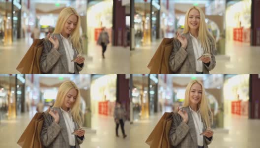 微笑优雅的年轻女子的肖像与长长的金发看着相机和使用手机站在购物中心的纸袋。高清在线视频素材下载