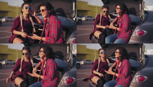 两个年轻漂亮的女人戴着墨镜坐在商场旁边停车场的敞篷后备箱里，用智能手机听着音乐。Slowmotion拍摄高清在线视频素材下载