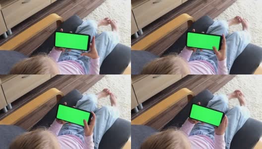 孩子坐在家里的椅子上使用智能手机，智能手机的绿色屏幕模型是水平景观模式。使用手机、浏览互联网、观看内容、视频、写博客、玩游戏高清在线视频素材下载