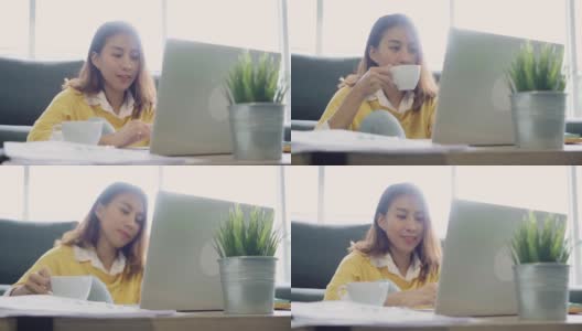 美丽的年轻微笑的亚洲女人工作在笔记本电脑和喝咖啡在家里的客厅。亚洲商业女性工作文件财务和计算器在她的家庭办公室。享受在家的时光。高清在线视频素材下载
