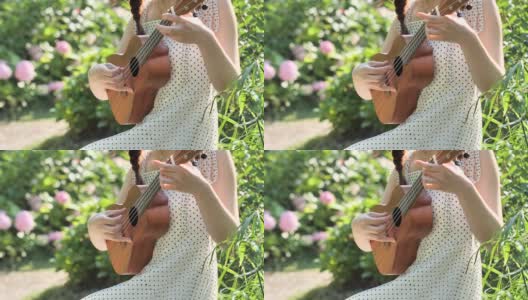 一个穿着裙子的女人的手拿着和弹奏四弦琴在花园与模糊的公园背景，4k视频，慢动作。高清在线视频素材下载