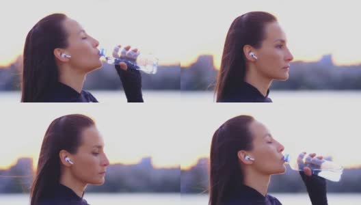 亚洲年轻漂亮的黑发女子穿着运动服，戴着无线耳机，从塑料瓶里喝水。她的头发在风中飘动。女孩自信地直视前方高清在线视频素材下载