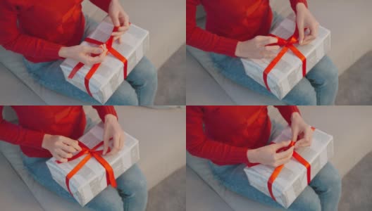 闭上女人的手用丝带打蝴蝶结为一个包装好的礼品盒。漂亮的女性穿着红衣服坐在客厅的沙发上，准备圣诞礼物庆祝圣诞传统派对高清在线视频素材下载