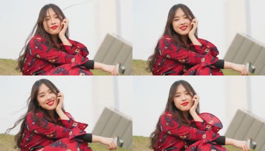 美丽的中国女孩穿着红衣服坐在草地上，享受微风吹拂她的长发，看着镜头，甜美的微笑，4k电影，慢镜头。高清在线视频素材下载