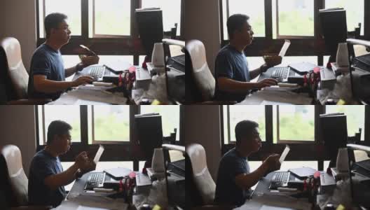 一个亚洲华人中年男子正在吃午餐，在他的家庭办公室工作的时候吃他的午餐高清在线视频素材下载