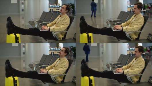 一个无聊的年轻人坐在机场，等待飞机起飞，使用着笔记本电脑。在笔记本电脑上打字，双腿放在黄色手提箱上。侧视图高清在线视频素材下载