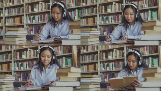 相当积极的亚洲青少年学生戴着耳机听音乐坐在大图书馆的桌子上拿着书和写总结高清在线视频素材下载