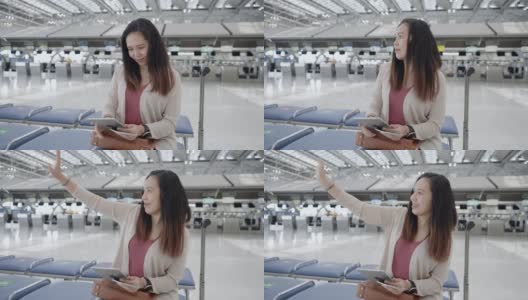一名亚洲女子身穿粉色t恤，带着行李、护照、机票等在值机柜台前的椅子上，用数码平板电脑检查航班时间，挥手微笑，在机场开始一段新的旅程，旅行，度假。高清在线视频素材下载
