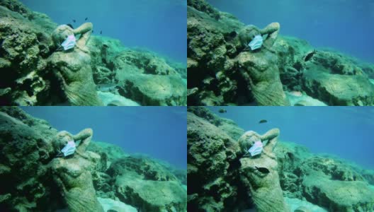 戴着医用面具的雕像躺在海底，旁边还有几条鱼。高清在线视频素材下载