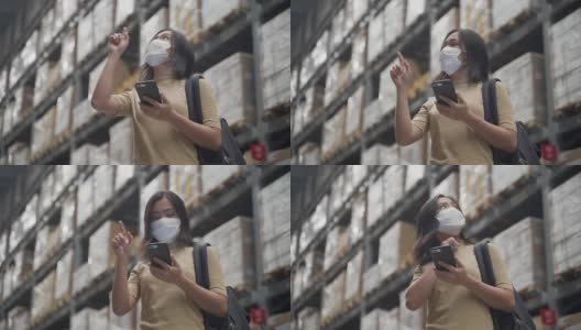亚洲妇女戴着面具站在仓库里，用智能手机查看价格。高清在线视频素材下载