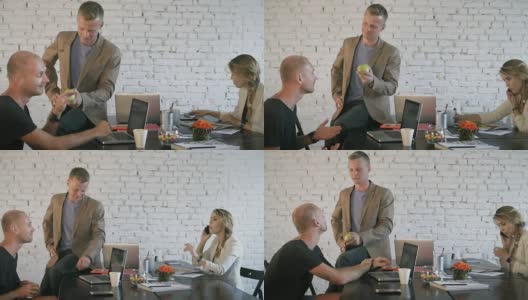 员工们在一个有创意的办公空间里相互交流。年轻的商人坐在桌子旁玩着苹果，和他的同事交流高清在线视频素材下载