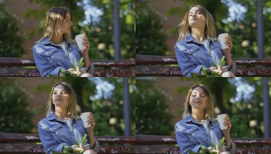 美丽的金发美女穿着牛仔夹克放松在公园长椅上喝咖啡高清在线视频素材下载
