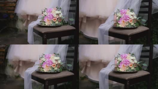 美丽的新娘花束在桌子上与美丽的婚纱和新娘饰品的背景。婚礼礼服在肩膀上，仪式前。美丽的婚礼花束和新娘的礼服高清在线视频素材下载