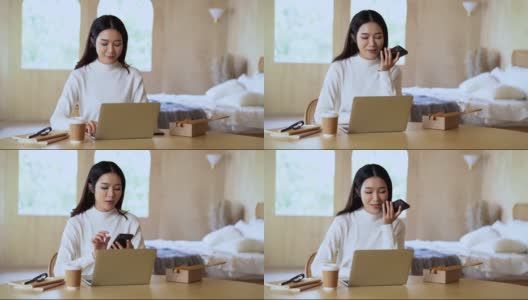美丽微笑的亚洲创意女性穿着毛衣，一边在家里用笔记本电脑工作一边讲手机。年轻的亚洲自由职业者妇女在家工作。小企业主。高清在线视频素材下载