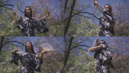 年轻苗条的日本女子手拿扇子慢动作跳舞。美丽的亚洲舞者穿着和服在公园里享受阳光明媚的春天。高清在线视频素材下载