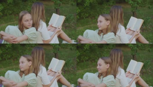 轻松的少女背靠背坐在绿色的草地上读书在暑假公园。一个年轻的女人在暑假的绿草地上看书。高清在线视频素材下载
