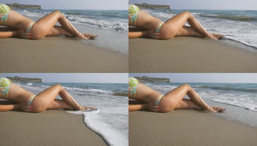 海浪冲刷着年轻女性的脚。难以辨认的迷人的女人在比基尼躺在海滩上。晒黑的女孩在美丽的海岸晒太阳。暑假或假日概念。关闭慢动作侧视图低角度视图高清在线视频素材下载