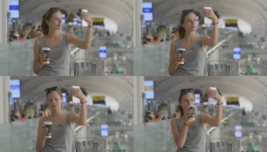 年轻的白人女性旅客在机场自拍。拿着手机和咖啡纸杯高清在线视频素材下载
