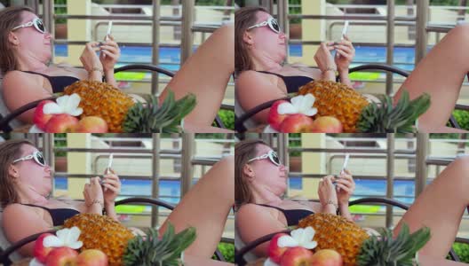 一名年轻女子戴着太阳镜躺在日光浴床上，在游泳池旁边用智能手机度假，前景是水果和鸡蛋花。3840 x2160高清在线视频素材下载