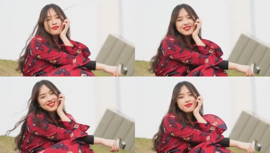 一名身穿红色连衣裙的美丽中国年轻女子坐在草地上，对着镜头微笑，享受微风吹拂她的黑色长发，4k视频，慢镜头。高清在线视频素材下载