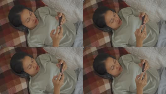 亚洲女人躺着，戴着无线耳机，用智能手机玩游戏高清在线视频素材下载