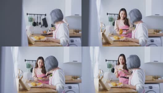 亚洲可爱的家庭，年轻的女儿看着老妈妈在厨房做饭。漂亮女性享受与年长的妈妈在家里餐桌上烤牛角面包的闲暇时光。活动的关系。高清在线视频素材下载