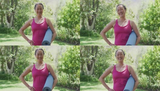 亚洲妇女的肖像，举着瑜伽垫微笑，站在公园高清在线视频素材下载