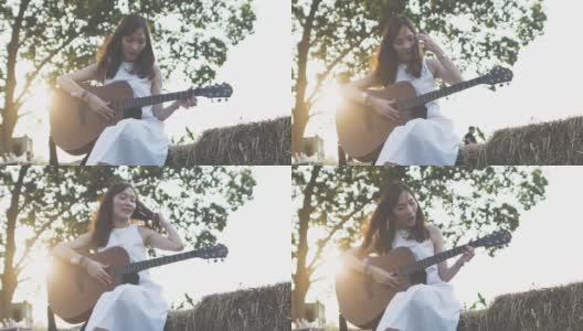 在清晨阳光的照射下，一名年轻美丽的亚洲女子穿着白衣在草堆上弹吉他。高清在线视频素材下载