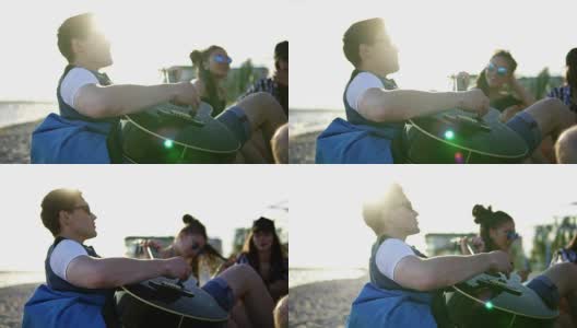 一个年轻人和一群朋友坐在沙滩上的安乐椅上弹吉他，在一个夏日傍晚的夕阳下唱歌。Slowmotion拍摄高清在线视频素材下载