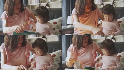 亚洲年轻美丽的母亲和她的小女儿一起在家里的沙发上看纪实书。他们指着书，看着它与幸福共处的时间。幸福的家庭的概念高清在线视频素材下载