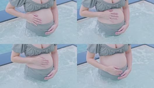 快乐孕妇站在水按摩按摩池抱着和抚摸她的大肚子在家里。年轻女子怀孕享受与未来生活放松在游泳池。怀孕的概念高清在线视频素材下载