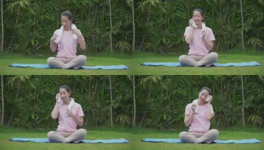 疲惫的年轻亚洲健身妇女练习瑜伽休息和擦汗毛巾坐在垫在公园早上。女性在户外的后院锻炼高清在线视频素材下载