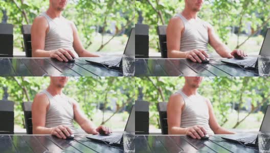 在户外庭院里用笔记本电脑工作——一个穿着t恤的男人坐在桌子旁的野营椅上。家庭办公室，远程工作，远程位置。使用键盘和鼠标，放松高清在线视频素材下载