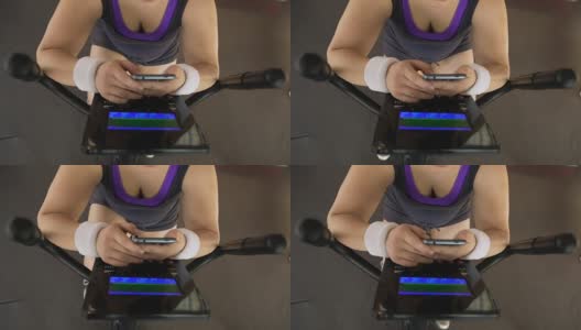 胖女人一边骑健身自行车一边刷手机，健身应用高清在线视频素材下载