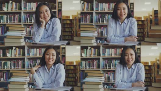 年轻美丽的亚洲女学生的肖像坐在桌子与图书馆的教科书看着相机。她满脸堆笑。高清在线视频素材下载