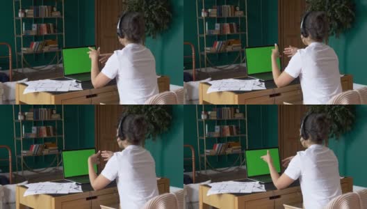 坐在椅子上的年轻女子在一台绿色模拟屏幕的笔记本电脑上工作。使用电脑的女孩，在线电话会议，与人培训，使用电脑。家中明亮的房间高清在线视频素材下载
