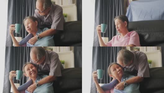 成熟的老人照顾女人，喝着热咖啡或茶坐在客厅的沙发上。年长的丈夫给妻子盖上毯子，并从后面拥抱她。快乐的退休生活。高清在线视频素材下载