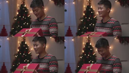 在装饰着圣诞树的充满节日气氛的客厅里，打开礼品盒的亚洲男子虽然感到不满和沮丧，但仍以礼貌的微笑表示感谢高清在线视频素材下载