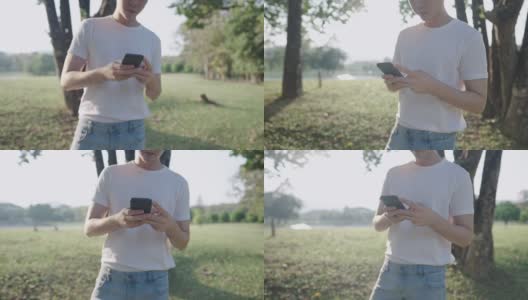 一名亚洲男子身穿白色休闲衬衫，一边走在绿色公园里一边使用智能手机，双手使用无线技术，放松休闲活动，无线上网，在树荫下等待高清在线视频素材下载