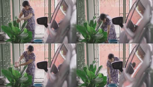 亚洲退休的老年妇女在她的家前面的一个小花园用回收塑料杯作为浇水罐浇灌植物叶子树，积极的老年生活方式的概念，侧视图高清在线视频素材下载