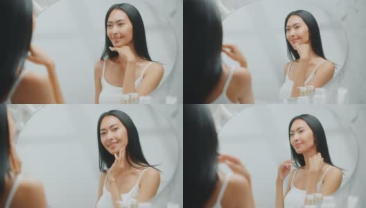 美丽的亚洲女人扮演和欣赏她完美柔软的皮肤和繁茂的黑发，看着相机和微笑在浴室的镜子。健康，自然美容，有机护肤品的概念高清在线视频素材下载