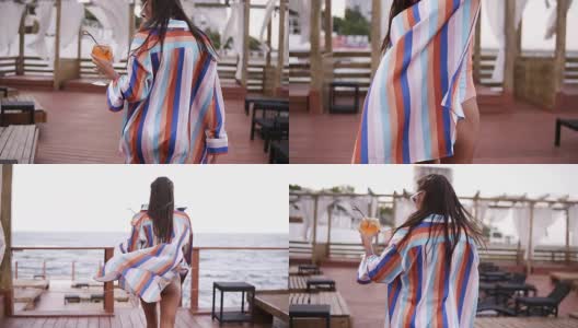 一个奢侈时尚的女人拿着一杯鸡尾酒，在海边的木制休息区走着彩色丝绸晨衣。夏季高清在线视频素材下载