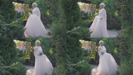 广角镜头华丽自信的资深新娘在白色优雅的婚纱旋转慢动作微笑看着镜头。有吸引力的白种人女人与灰色的头发摆姿势在户外阳光。高清在线视频素材下载