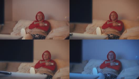 穿着红色运动衫，头戴兜帽的男人晚上坐在沙发上看电视，摄像机在移动高清在线视频素材下载