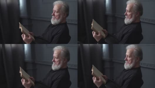 跟踪拍摄的深思熟虑的白发老人阅读旧纸质书在黑暗的图书馆房间。高清在线视频素材下载
