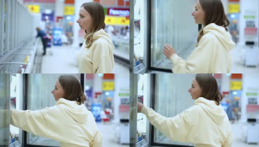 一个女人在超市买冷冻食品。一个漂亮的年轻女人站在冰箱旁边挑选鱼。在超市买食物高清在线视频素材下载