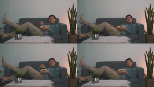 一名亚洲男子在晚上看电影时斜靠在沙发上吃宵夜。高清在线视频素材下载