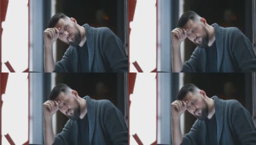 一个英俊的胡子男子的近距离肖像坐在晚上靠窗的街道上，他的头支撑在他的手，并看着沉思低头在他的笔记本电脑的屏幕高清在线视频素材下载