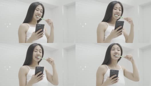 这是一个美丽的20多岁的亚洲女性早上对着浴室镜子刷牙，兴奋地微笑着查看手机短信和邮件的照片高清在线视频素材下载